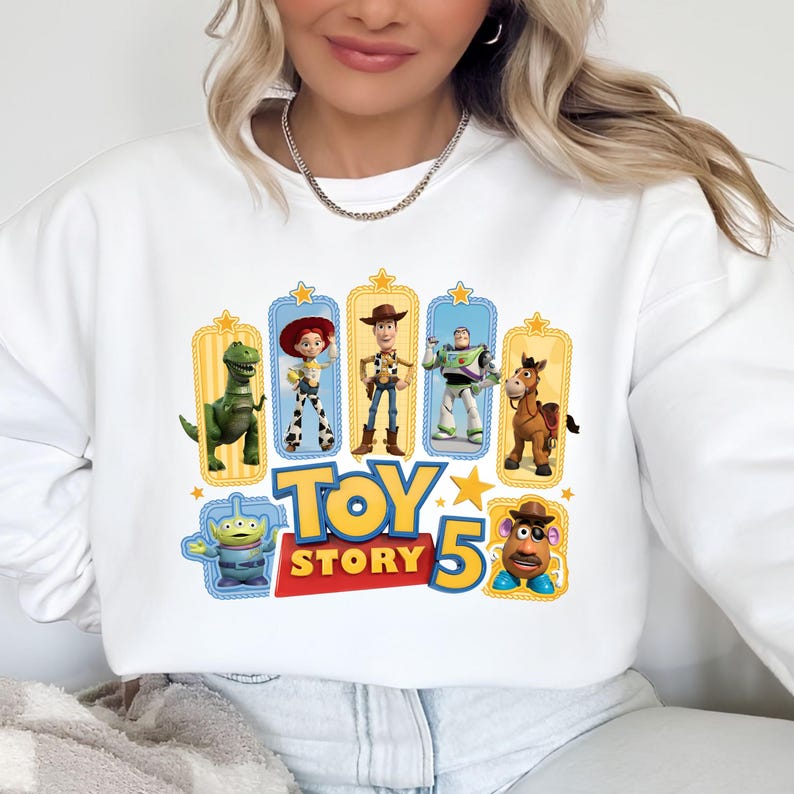 Op de afbeelding: Een wit sweatshirt met lange mouwen en een kleurrijke grafische print van Toy Story-personages, waaronder Rex, Jessie, Woody, Buzz Lightyear, Bullseye, een alien en Mr. Potato Head, met de tekst Toy Story 5 in het geel en rood.
