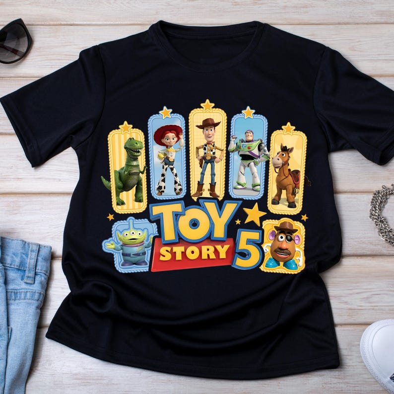 Op de afbeelding: Een zwart t-shirt met korte mouwen en een grafische print van Toy Story-personages zoals Rex, Jessie, Woody, Buzz Lightyear, Bullseye, een alien en Mr. Potato Head, met de tekst Toy Story 5 in grote gele en rode letters.
