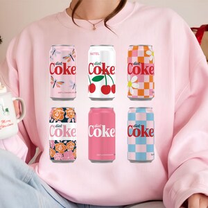 May include: A light pink sweatshirt featuring six Diet Coke cans with various designs. The cans showcase floral, checkered, and dragonfly patterns, with the Diet Coke logo in red. The sweatshirt is a solid light pink color.