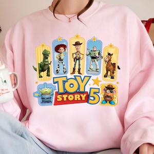 Op de afbeelding: Een lichtroze sweater met ronde hals en een grafische print van Toy Story-personages, waaronder Rex, Jessie, Woody, Buzz Lightyear, Bullseye, een alien en Mr. Potato Head, met de tekst Toy Story 5 in grote gele en rode letters.