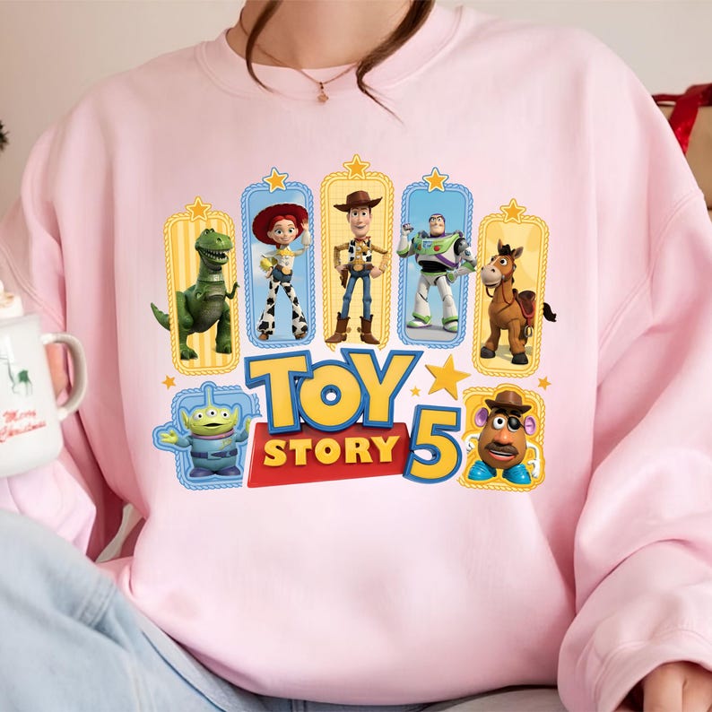 Op de afbeelding: Een lichtroze sweater met ronde hals en een grafische print van Toy Story-personages, waaronder Rex, Jessie, Woody, Buzz Lightyear, Bullseye, een alien en Mr. Potato Head, met de tekst Toy Story 5 in grote gele en rode letters.