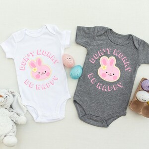 May include: Two baby onesies, one white and one gray, with a pink bunny graphic and the text "Don't Worry Be Happy." Easter eggs and a stuffed bunny are also in the image. The onesies are short-sleeved and have a round neckline.