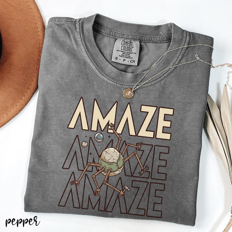 May include: A gray Comfort Colors t-shirt with the word "AMAZE" in a light tan color, repeated three times with a robot-like creature graphic. A silver necklace with a pendant is on the shirt.
