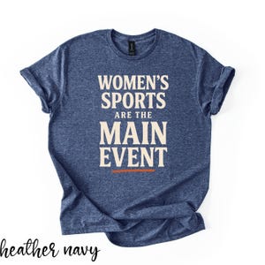 May include: A heather navy blue short-sleeve t-shirt featuring the cream-colored text "WOMEN'S SPORTS ARE THE MAIN EVENT" with a thin orange horizontal line underneath the text. The shirt has a crew neckline and a standard fit.