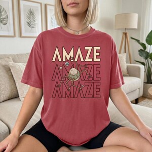 May include: A red t-shirt with the word "AMAZE" stacked three times in a retro font. The middle "AMAZE" features a graphic of a robotic insect with a spherical head and multiple legs. The shirt is a solid red color.