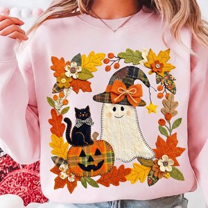 Puede incluir: Una sudadera rosa claro con un gráfico central de un fantasma blanco con sombrero de bruja a cuadros y lazo naranja, un gato negro con lazo a cuadros sobre una calabaza a cuadros, rodeado por una corona de hojas de otoño coloridas.