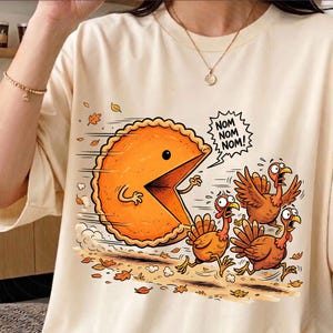May include: A cream-colored t-shirt featuring a graphic of a large, orange, circular pie with a mouth open, chasing three brown turkeys. The pie has a speech bubble that says "NOM NOM NOM!" and is surrounded by scattered autumn leaves.