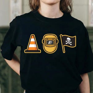 K&ouml;nnte beinhalten: Schwarzes T-Shirt mit drei Cartoon-Grafiken: einem orange-wei&szlig;en Verkehrskegel, einer gelben Schwei&szlig;maske und einer schwarzen Flagge mit Totenkopf und gekreuzten Knochen. Das Wort "AOP" ist impliziert.