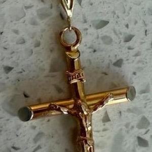 May include: Gold-toned crucifix pendant on a matching chain. The cross features a detailed depiction of Jesus Christ. The chain has a woven design. The pendant and chain are displayed against a light-colored, speckled background.