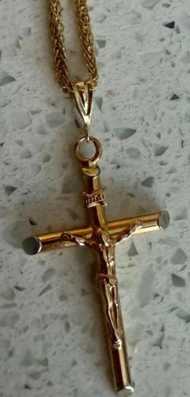 May include: Gold-toned crucifix pendant on a matching chain. The cross features a detailed depiction of Jesus Christ. The chain has a woven design. The pendant and chain are displayed against a light-colored, speckled background.