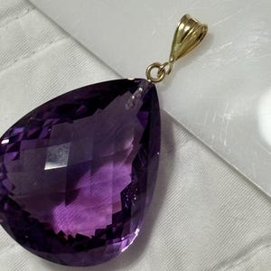 May include: A faceted, pear-shaped amethyst pendant with a gold bail. The deep purple gemstone has a polished surface and is suspended from a small, gold-colored loop. The pendant is set against a white background.