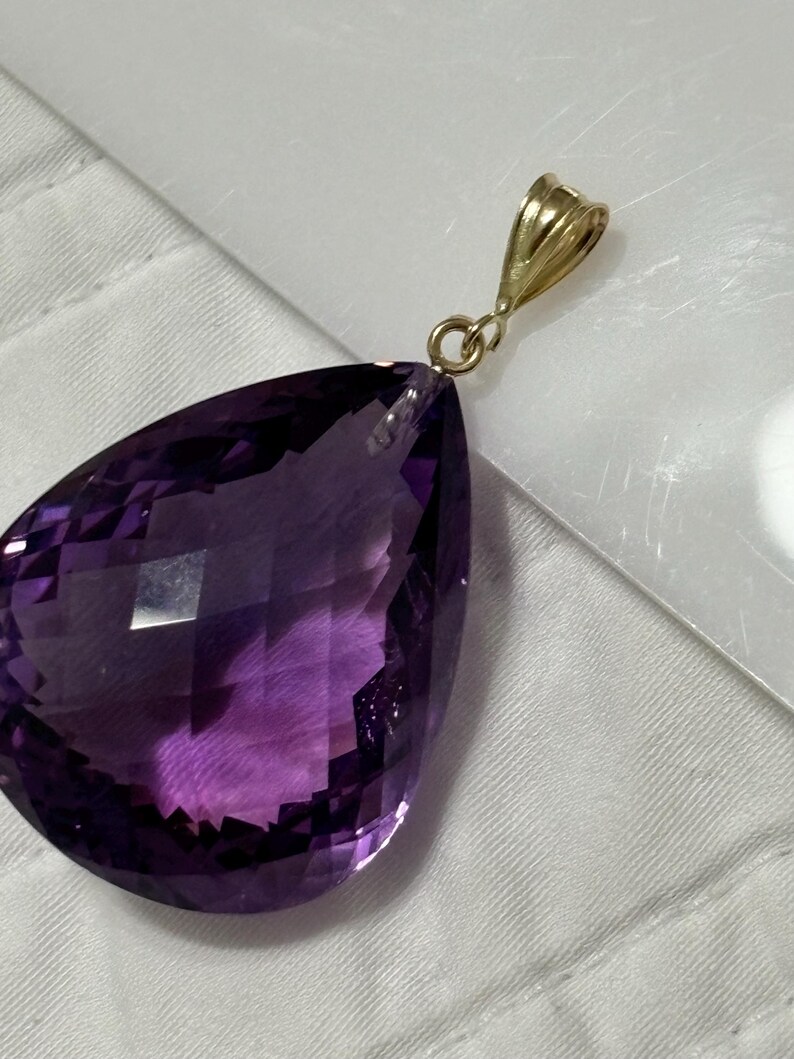 May include: A faceted, pear-shaped amethyst pendant with a gold bail. The deep purple gemstone has a polished surface and is suspended from a small, gold-colored loop. The pendant is set against a white background.