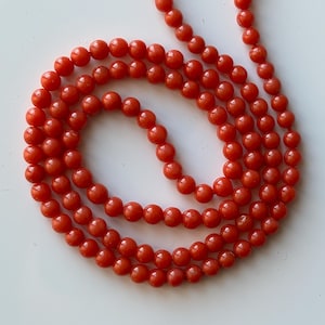 May include: A necklace of small, round, bright red beads. The beads are strung together, forming multiple strands that lay in a curved pattern. The beads have a smooth, glossy finish, and the overall effect is one of simple elegance.
