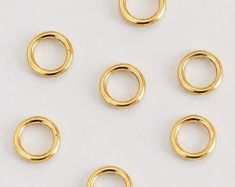 May include: A collection of seven circular gold-toned metal jump rings arranged in a scattered pattern. Each ring features a smooth, polished surface with a visible seam where the ends meet, creating a consistent metallic appearance throughout.