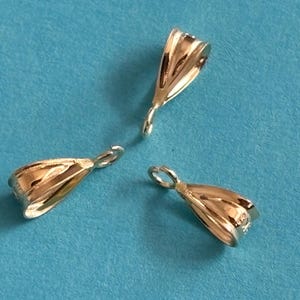 May include: Three silver-colored jewelry bails, each with a loop at the top and a curved, open design. The bails are displayed against a solid blue background, creating a contrast that highlights their metallic sheen.