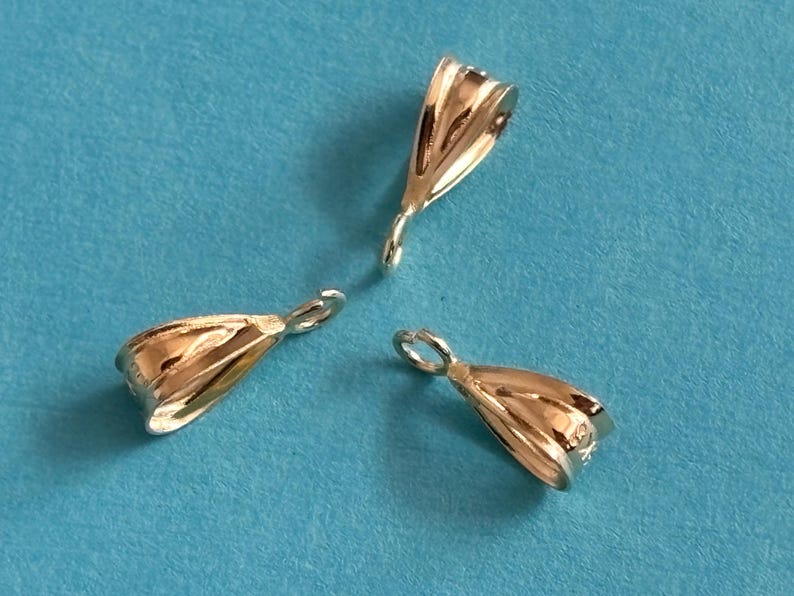 May include: Three silver-colored jewelry bails, each with a loop at the top and a curved, open design. The bails are displayed against a solid blue background, creating a contrast that highlights their metallic sheen.