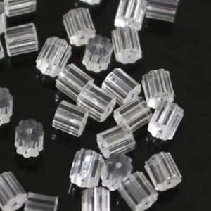 May include: A collection of clear, plastic earring backs. The backs are cylindrical with ribbed sides and a flower-shaped top. They are scattered on a dark surface, creating a pattern of small, transparent shapes.
