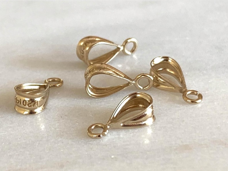 May include: A collection of gold-colored pendant bails. The bails have a teardrop shape with a small loop at the top and a wider opening at the bottom. One bail has the number "2019" engraved on it.