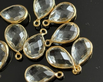 May include: A group of clear faceted teardrop-shaped glass beads with gold-toned metal loops. The beads are arranged on a black surface.