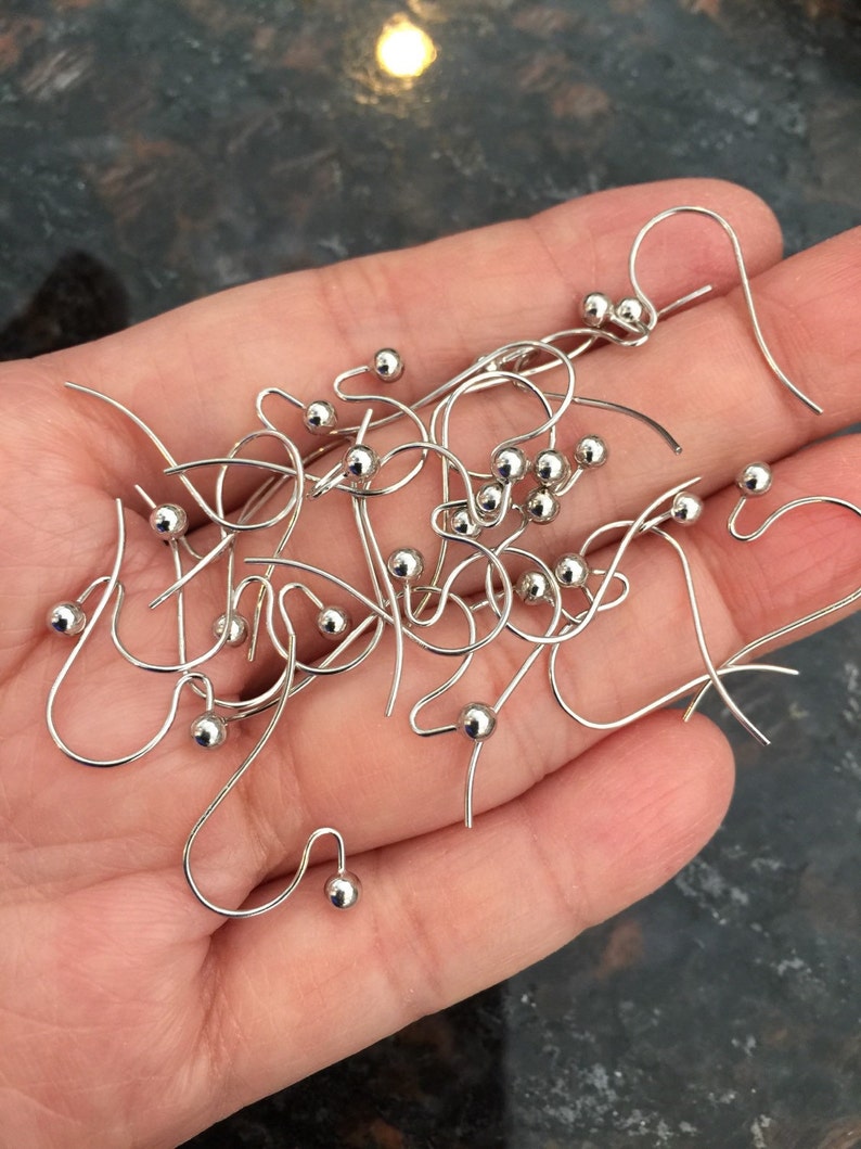 Puede incluir: Primer plano de un mont&oacute;n de ganchos para pendientes de plata con peque&ntilde;as bolas redondas en el extremo. Los ganchos son de metal y est&aacute;n dise&ntilde;ados para sujetar pendientes.