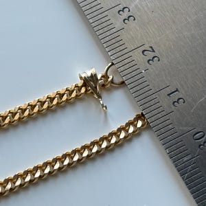 May include: Close-up of a gold-colored curb chain necklace with a pendant. A metal ruler is placed next to the chain, showing measurements. The chain is a warm gold color and has a textured, rounded design.