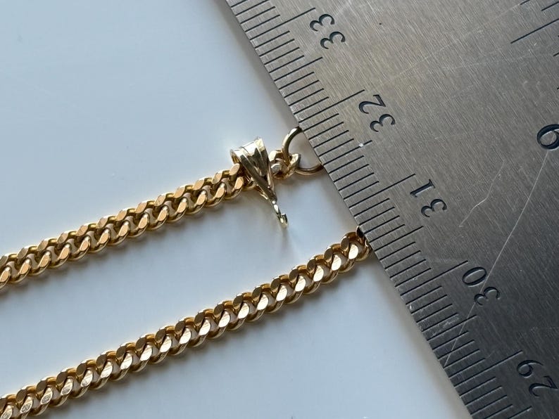 May include: Close-up of a gold-colored curb chain necklace with a pendant. A metal ruler is placed next to the chain, showing measurements. The chain is a warm gold color and has a textured, rounded design.