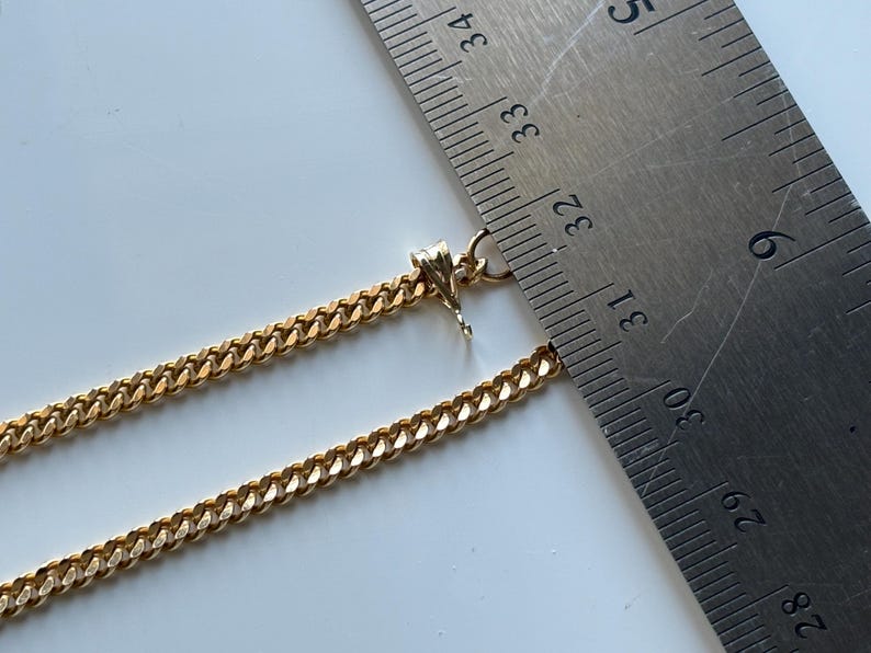 May include: A gold-colored curb chain necklace with a lobster clasp. The necklace is measured with a metal ruler, showing a length of approximately 12.5 inches. The chain links are closely linked together.