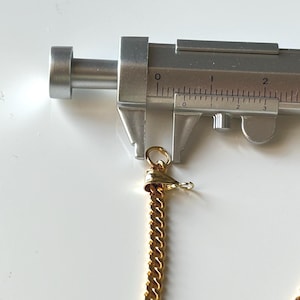 May include: A gold-colored chain necklace with a lobster clasp is measured with a silver caliper. The chain is a curb style, and the clasp is attached to a small gold-colored ring. The caliper shows a measurement of approximately 0.5 inches.