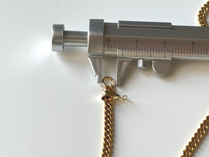 May include: A gold-colored chain necklace with a lobster clasp is measured with a silver caliper. The chain is a curb style, and the clasp is attached to a small gold-colored ring. The caliper shows a measurement of approximately 0.5 inches.