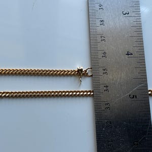 May include: A gold-colored chain necklace with a curb link design. The necklace is measured with a ruler, showing a length of approximately 14 inches. The chain is displayed on a white surface.