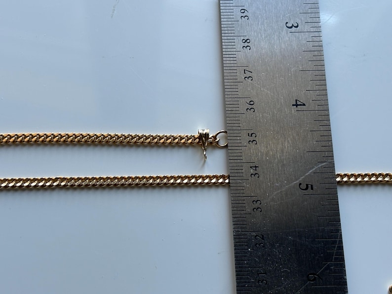 May include: A gold-colored chain necklace with a curb link design. The necklace is measured with a ruler, showing a length of approximately 14 inches. The chain is displayed on a white surface.
