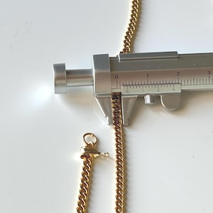 May include: A gold-colored curb chain necklace is measured with a silver caliper. The chain is approximately 0.1 inches thick. A gold clasp is visible at the end of a separate chain segment. The background is white.