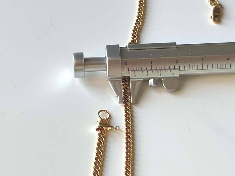 May include: A gold-colored curb chain necklace is measured with a silver caliper. The chain is approximately 0.1 inches thick. A gold clasp is visible at the end of a separate chain segment. The background is white.