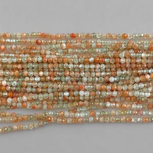 Może przedstawiać: Kolekcja fasetowanych okrągłych koralików ułożonych w wiele poziomych pasm. Koraliki charakteryzują się mieszanką półprzezroczystej jasnej zieleni i nieprzezroczystej pomarańczowej czerwieni, tworząc zróżnicowany wygląd na całej długości pasm.