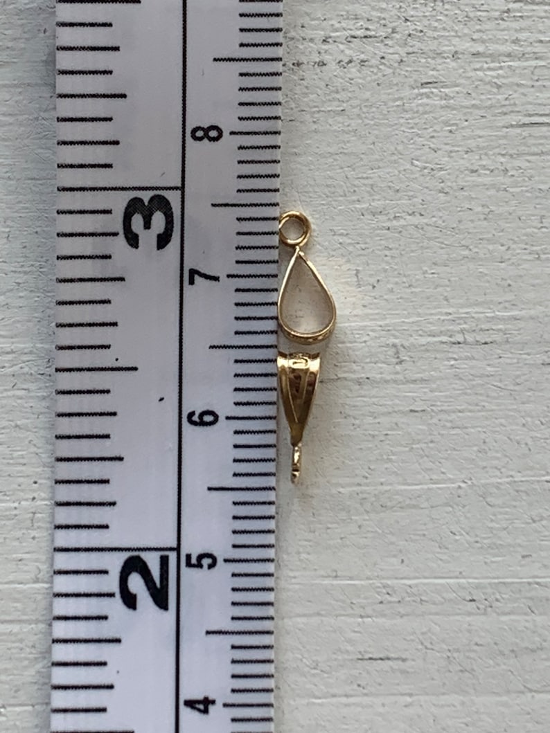 May include: A gold-colored jewelry bail with a teardrop-shaped setting. The bail measures approximately 0.75 inches in length. The bail is designed to hold a pendant or charm. The background is a light-colored wooden surface.