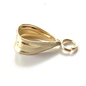 May include: A gold-colored pendant bail with a teardrop shape and a small attached jump ring. The bail has a textured surface and is designed to hold a pendant. The bail is made of a shiny, reflective metal.