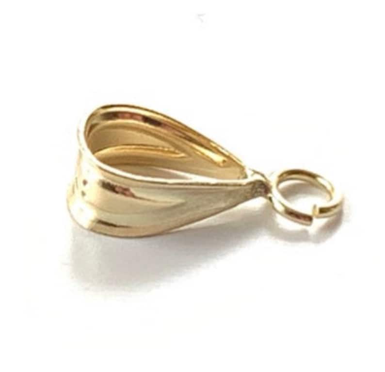 May include: A gold-colored pendant bail with a teardrop shape and a small attached jump ring. The bail has a textured surface and is designed to hold a pendant. The bail is made of a shiny, reflective metal.