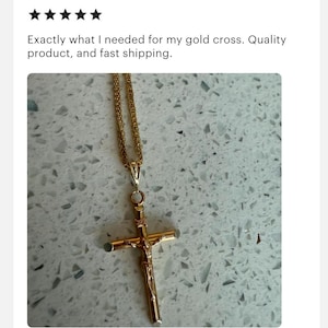 May include: A gold-colored pendant necklace featuring a crucifix design. The cross has a detailed depiction of Jesus. The necklace chain is also gold-colored. The image is set against a light-colored, speckled background.