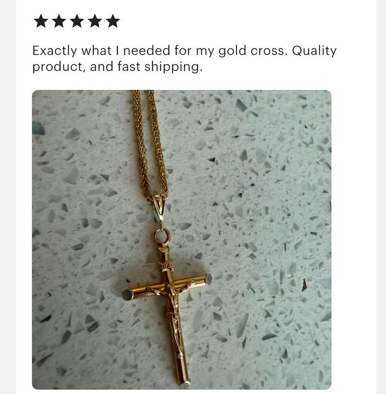 May include: A gold-colored pendant necklace featuring a crucifix design. The cross has a detailed depiction of Jesus. The necklace chain is also gold-colored. The image is set against a light-colored, speckled background.