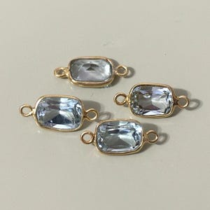 May include: Four rectangular, light blue gemstone charms with gold-colored bezels and small loops on each end. The gemstones have a faceted appearance. The charms are displayed on a light beige surface.