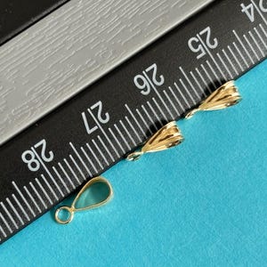 May include: Close-up of three gold-colored jewelry findings on a blue surface. Two findings have a textured, folded design, and the third is a teardrop shape with a loop. A black and gray ruler is in the background.
