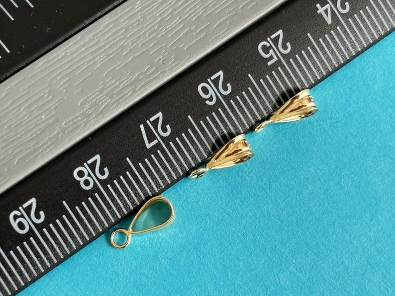 May include: Close-up of three gold-colored jewelry findings on a blue surface. Two findings have a textured, folded design, and the third is a teardrop shape with a loop. A black and gray ruler is in the background.