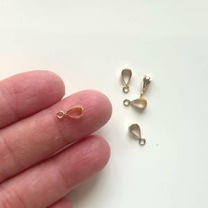 May include: Several small, gold-colored teardrop-shaped jewelry findings. One is held between fingers, while others are scattered on a white surface. Each finding has a small loop at the top and a teardrop-shaped body.