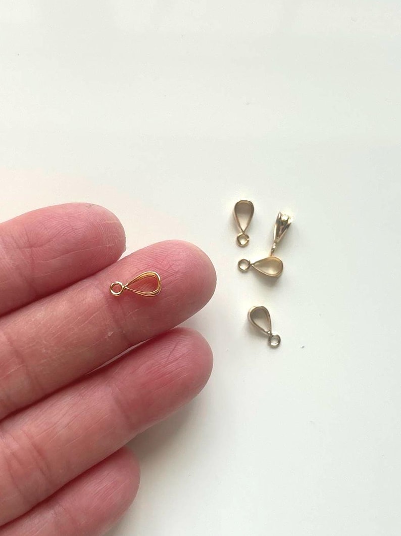 May include: Several small, gold-colored teardrop-shaped jewelry findings. One is held between fingers, while others are scattered on a white surface. Each finding has a small loop at the top and a teardrop-shaped body.