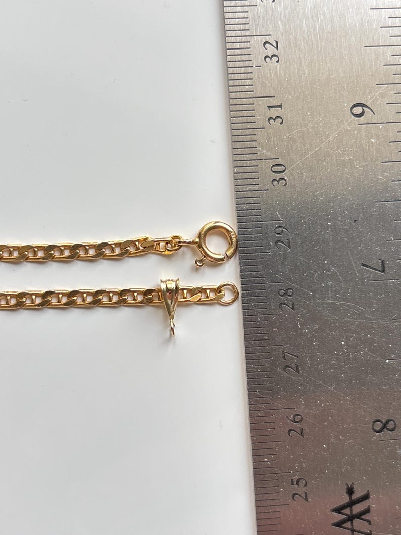 May include: A gold-colored chain necklace with a lobster clasp and pendant bail. The chain is laid out next to a ruler, showing its length. The necklace is on a white surface.