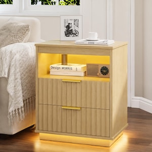 May include: A light oak nightstand with two drawers and a shelf. The nightstand has a ribbed texture on the drawer fronts and gold-colored handles. The shelf contains books, a small radio, and a decorative item. The nightstand has built-in lighting.