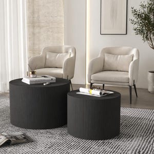 May include: Two black, cylindrical coffee tables with vertical ribbed detailing. The tables are different sizes and are in front of two cream-colored armchairs. A rug with a black and white pattern is on the floor.