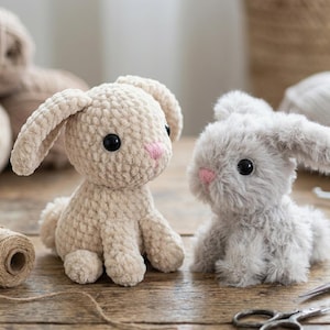 May include: Two plush bunny rabbits. One is a light beige color, crocheted with black eyes and a pink nose. The other is a soft gray with black eyes and a pink nose. Both have long ears and are sitting upright.