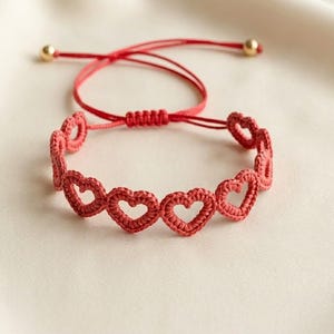 May include: A red crochet bracelet featuring a series of heart shapes. The bracelet is adjustable with a sliding knot and has gold-colored beads at the ends. The bracelet is displayed on a cream-colored surface.