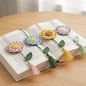 Puede incluir: Marcadores de libros de flores de ganchillo en rosa, azul, amarillo y morado, con hojas y borlas verdes. Los marcadores están colocados en las páginas de un libro abierto, sobre una mesa de madera. Hecho a mano.